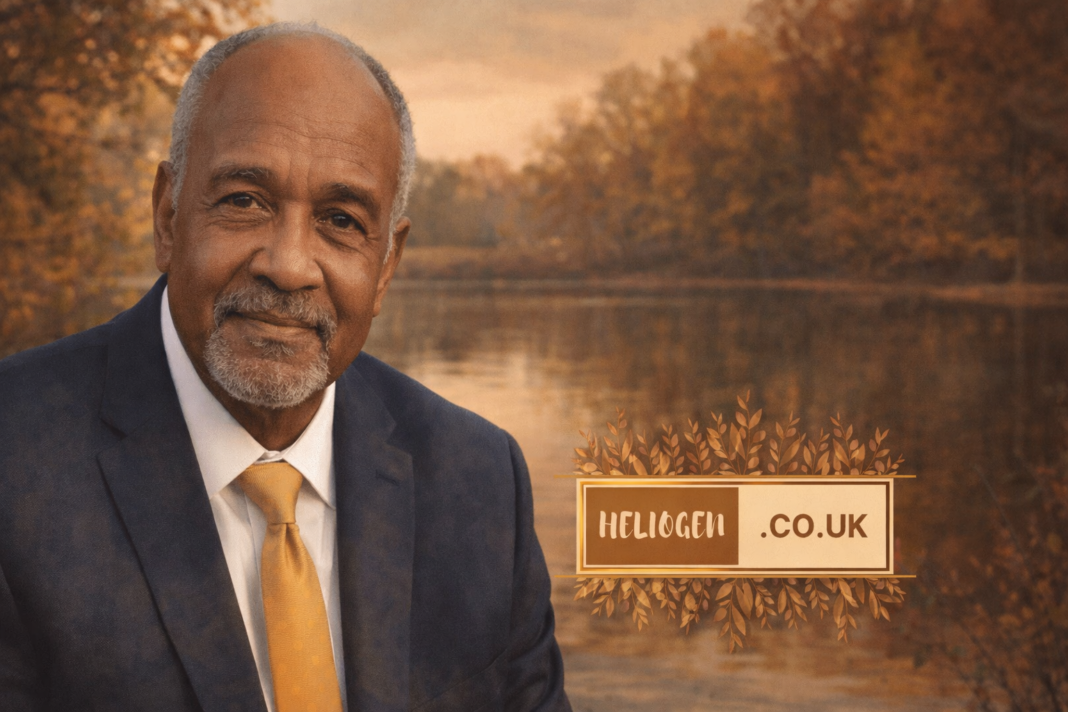Portrait of Claude Edward Elkins Jr. in a suit and gold tie, standing by a calm river with autumn trees in the background.